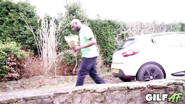 Bald black man holding flowers near white Renault by hedge