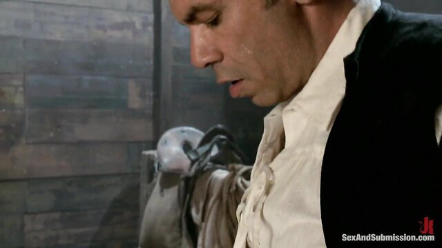 Man in period costume sweating, masked figure behind in slave auction scene