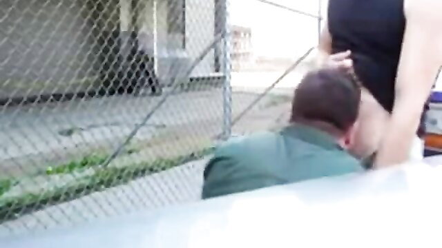 Woman giving outdoor blowjob to man on lunch break by chainlink fence