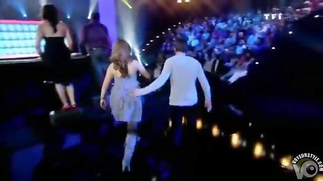 Busty goddess in short dress holds hand on TV quiz stage with crowd