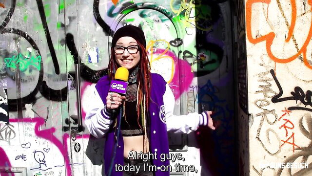 Punk girl with dreads and glasses poses with mic in varsity jacket