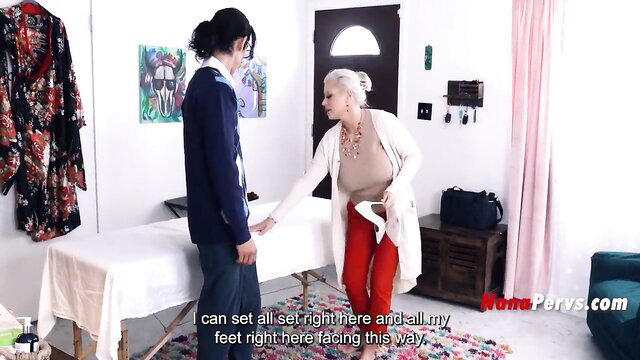 Blonde mature nana in red pants guides young guy at massage table