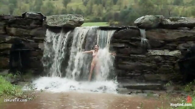 Nude brunette stands under outdoor waterfall, wet body glistening