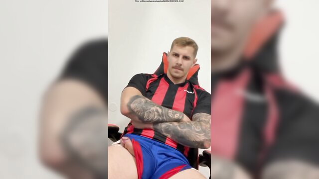 Tattooed blond bull in tight black red soccer jersey and blue shorts, muscular arms crossed