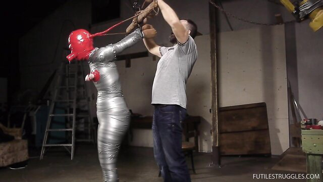 Diamondly mummified in silver tape, red hood, man hoists her arms in bondage