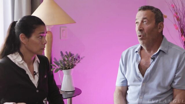 Asian woman in office attire interviews older man in pink studio