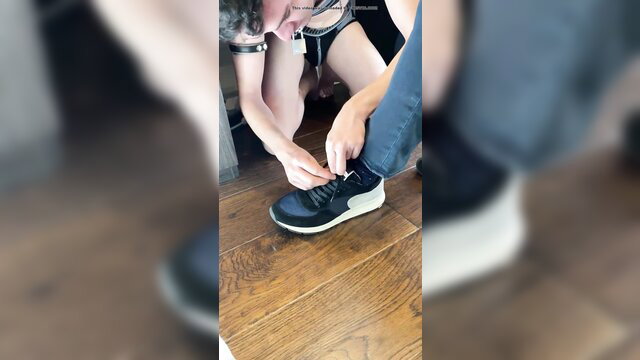 Slave boy kneels serving master, lacing black sneakers in BDSM scene