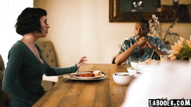 Curvy brunette Olive in green dress chats with Isiah at dinner table