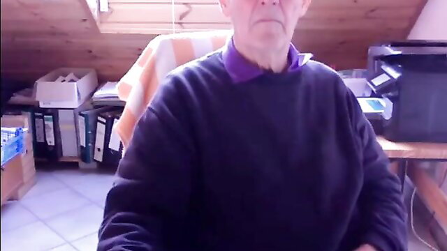 74yo German grandpa in purple shirt sits smiling in home office