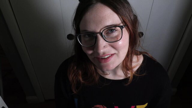 Smiling redhead MILF in glasses with cat ears, black top, low light tease