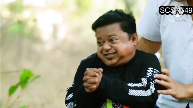 Smiling Indian dwarf man in black hoodie praying outdoors, hot Desi woman behind