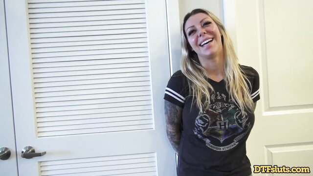 Karma Rx smiles in Slytherin tee, tattooed arm by bathroom door