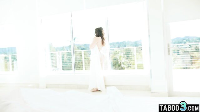 Teenage brunette in white dress stands nude from behind at window