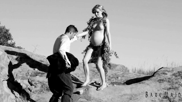 Pregnant blonde KellyGlass in grass skirt helped by man on rocks in Bare Maidens