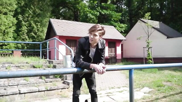 Slim twink in black leather jacket leans on railing in outdoor scene