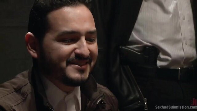 Dark-haired man in leather jacket smirks in dimly lit BDSM scene