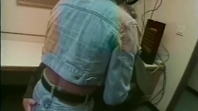 Blonde in denim jacket and jeans bends over desk in erotic pose