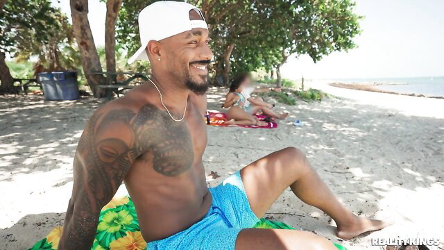 Tattooed black hunk in blue swim trunks on beach blanket, Reality Kings