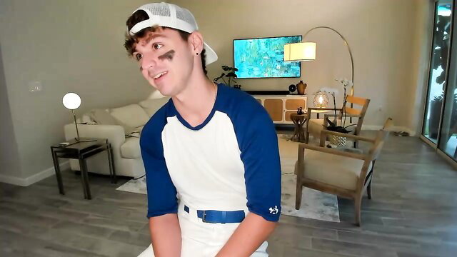 Cute twink in baseball cap smiles playfully on sofa in gay oral tease