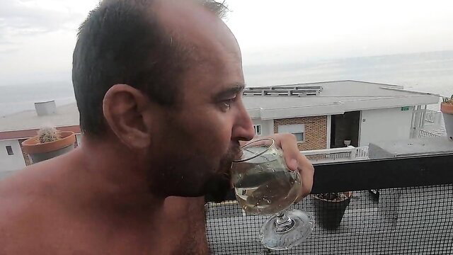 Bald muscular man sips pee from wine glass on balcony
