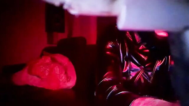 Crystalbitchx in shiny latex jacket next to red heart pillow, seductive red lighting