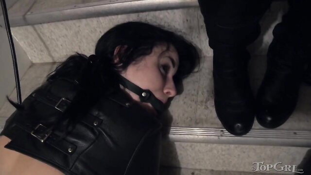 Bound brunette in leather neck brace lies helpless on stairs in lesbian fetish scene