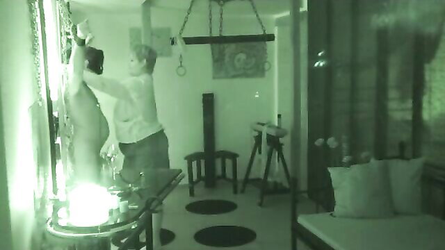 Nude blonde thrall bound at BDSM cross in green-lit dungeon