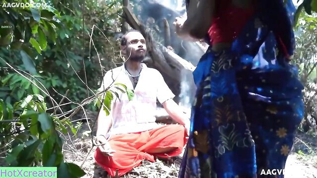 Naughty Indian tantrik in white kurta orange dhoti sits meditating as saree woman approaches in jungle