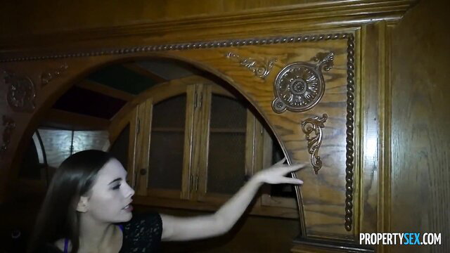 Petite brunette Skye West touching ornate wood cabinet in dim kitchen
