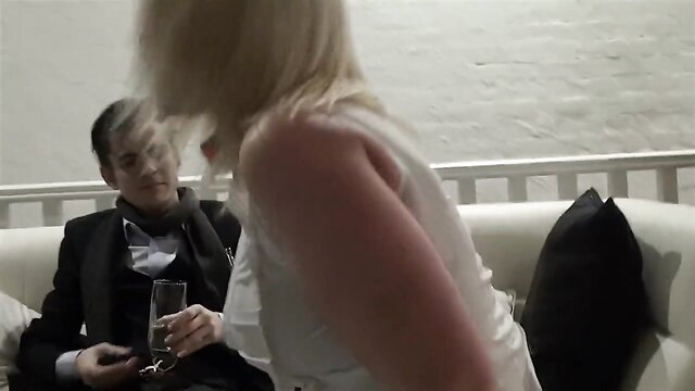 Blonde woman in white dress teases suited man during collar training session