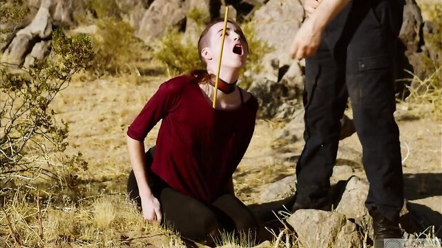 Bald woman kneels outdoors, mouth open wide with rod, man in tactical gear above