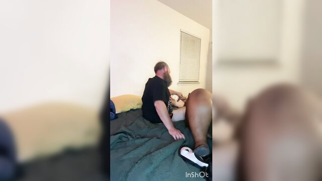 Bearded man sits on bed with legs spread, hand on thigh in bedroom