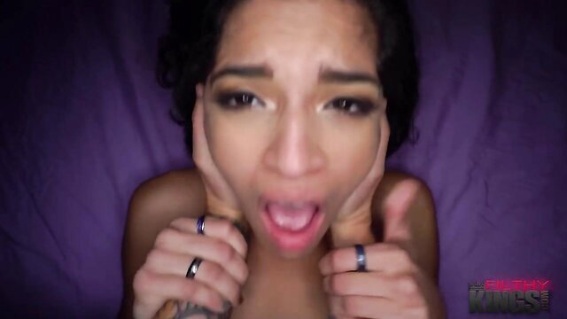 Tyler Steel, a Latina woman, lying on a purple bed, looking at the camera with her tongue out