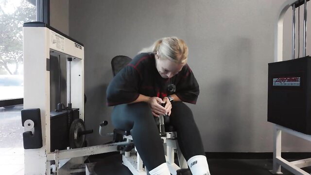 Blonde fitness girl in tight leggings seated on gym leg machine