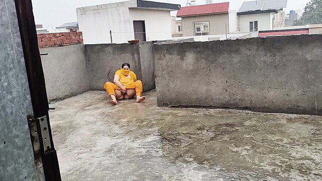 Mature Pakistani Aunty in yellow outfit sits on rooftop enjoying rain, ample melons booty
