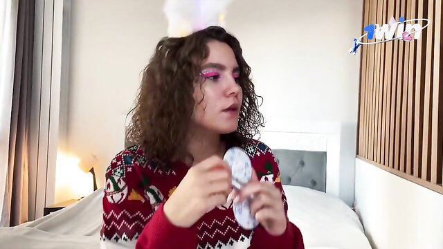Curly-haired brunette in Santa hat and sweater holds toy on bed
