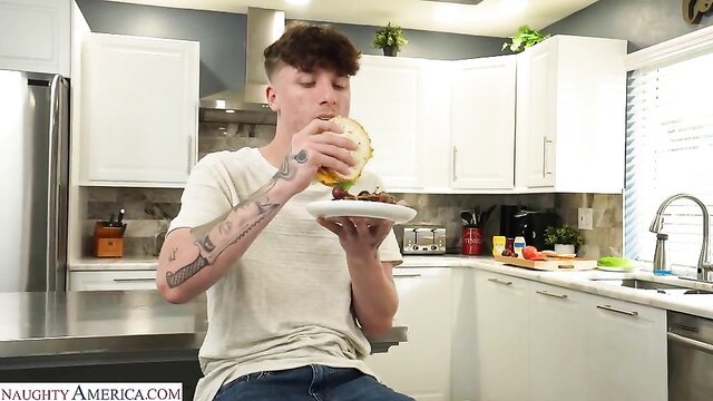 Tattooed white guy with curly hair eating burger in kitchen