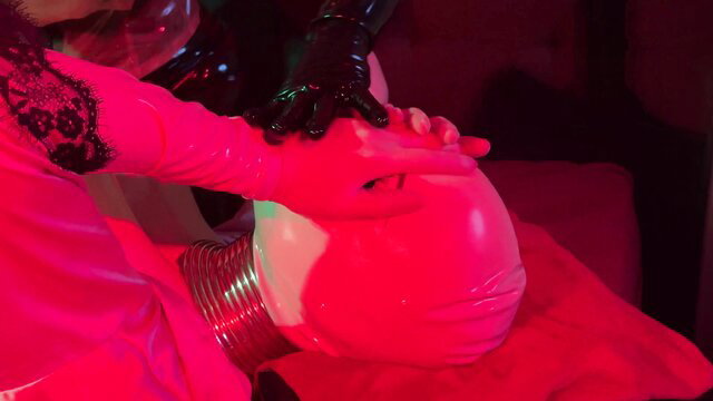 Rubber fetish domme in latex gloves fits red latex hood on sub's neck corset