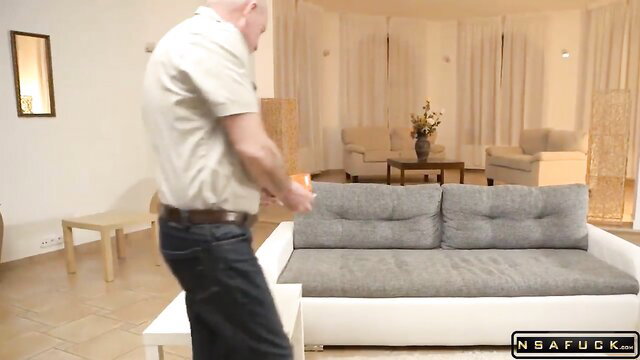 Bald older man in jeans holding orange object near gray sofa