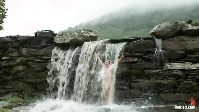 Nude woman with arms raised under cascading waterfall, wet outdoor fantasy