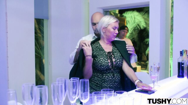 Blonde MILF in green dress at bar with man embracing her from behind