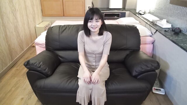 Cute Asian amateur sits smiling in beige dress on couch