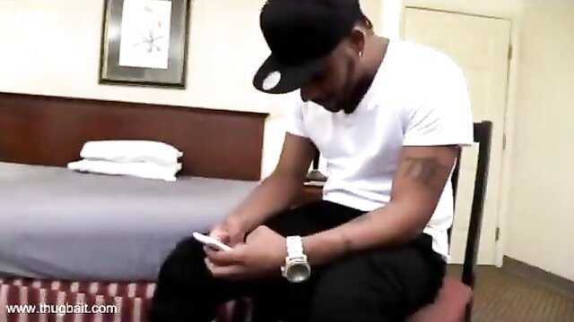 Black guy in white tee and cap sits on chair holding phone in hotel room
