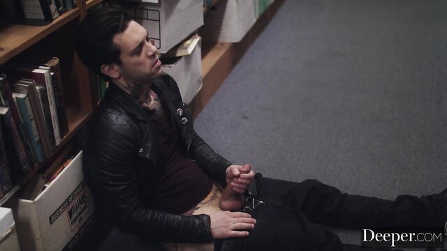 Tattooed man in leather jacket sits on floor holding cock in library