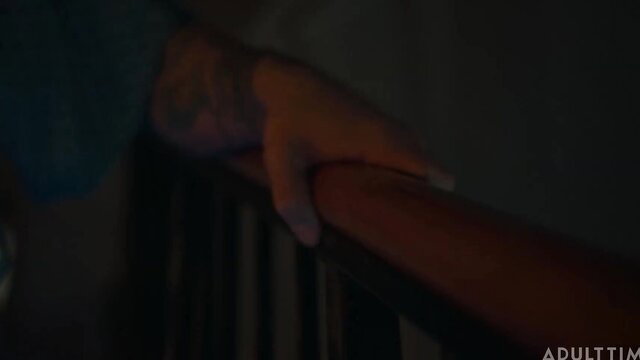 Tattooed hand gripping wooden railing in dim-lit adult scene