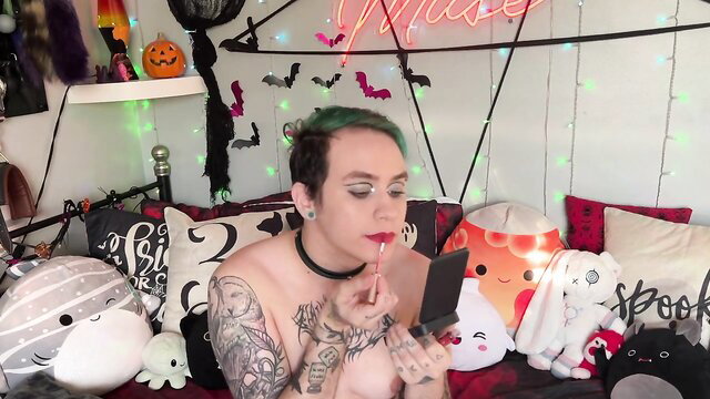 Tattooed goth girl with green hair applies red lipstick topless