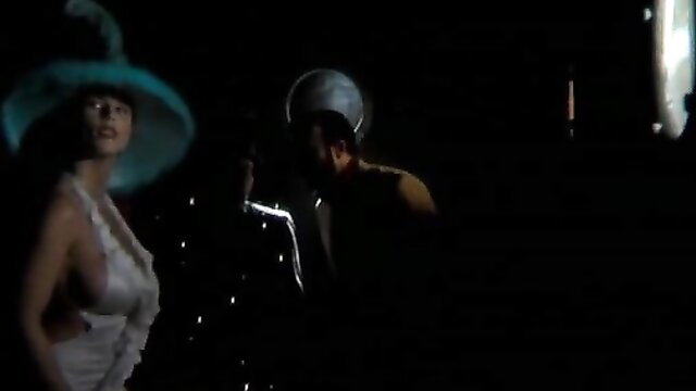 Sexy brunette in sheer dress and hat with suited man in dark scene