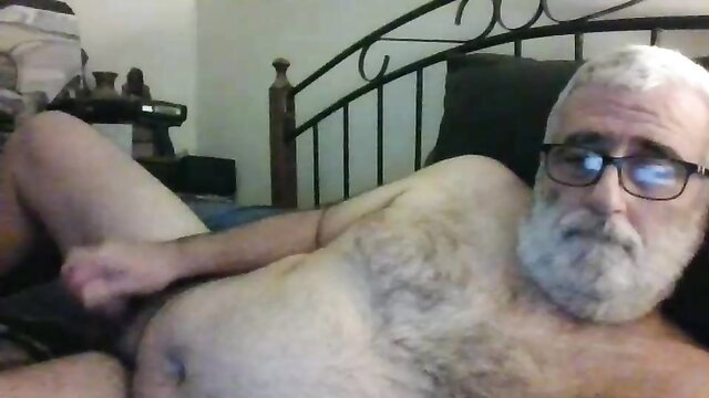 Hairy older white man with glasses lying nude stroking cock on bed