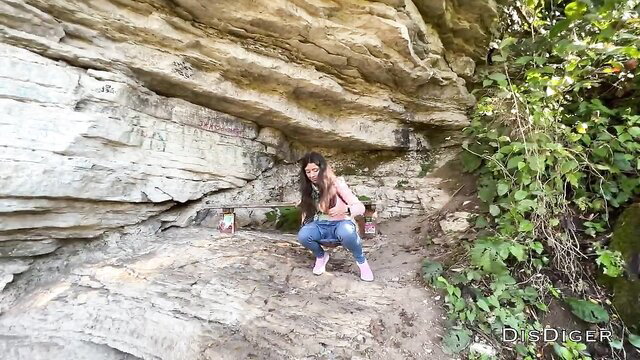 Sexy brunette squats in forest cave, tight jeans hug curves, outdoor tease