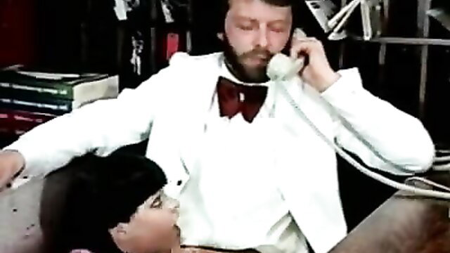 Retro Hotel Harlots Color Climax: Bearded man in white suit phones while woman kneels under desk